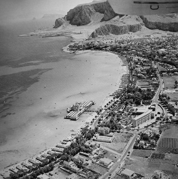 024_mondello.jpg - Un’altra veduta aerea che mostra Valdesi già abbastanza urbanizzata, mentre l’Addaura è ancora una landa desolata. La parte scurea del mare è una foresta di poseidonia, alga che ospita vita di pesci e crostacei e a cui è attribiito il merito della resistenza della spiaggia alle pericolose erosioni provocate dalle mareggiate. I lotti di terra, confinanti con la strada del lungomare, si preparano ad essere occupati dalle ville, stranamente costruite a limite verso il mare e con i giardini a monte.